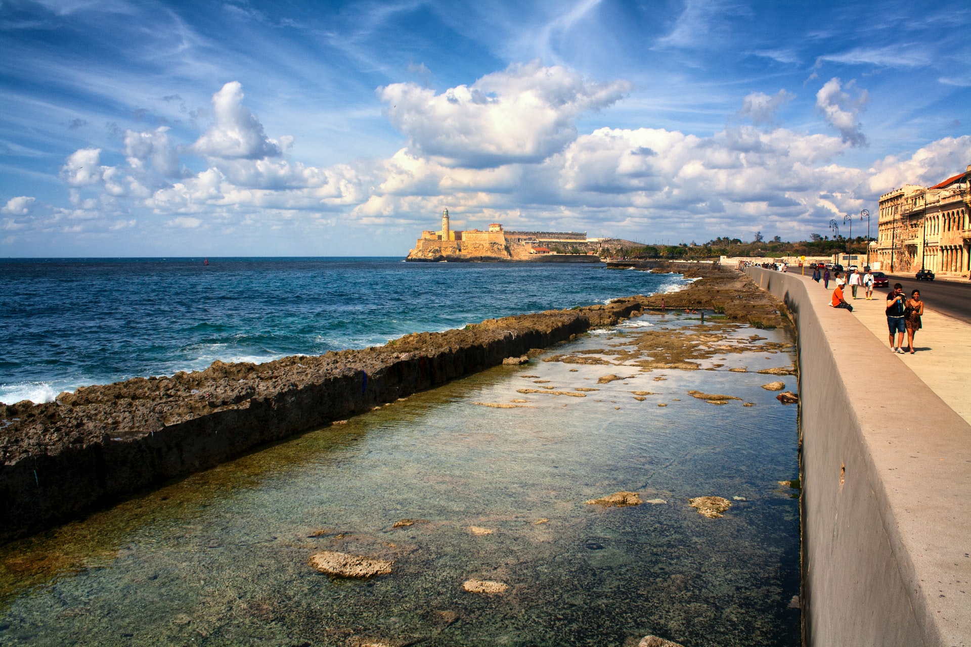 People walking along a promenade next to the sea with an old fortress on the coast in the background