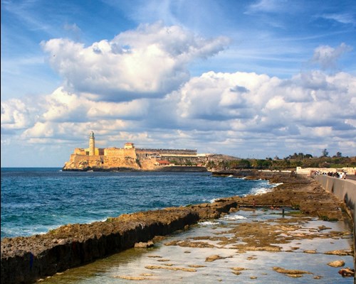 People walking along a promenade next to the sea with an old fortress on the coast in the background