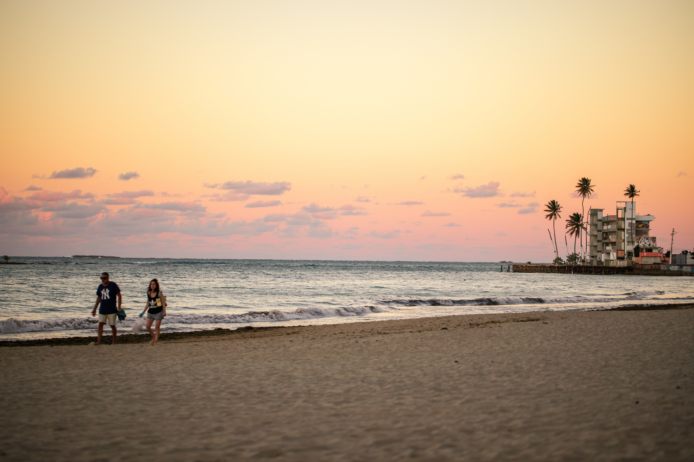 Peachy purple sunset with two people walking on the beach and city area in the distance