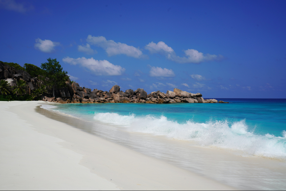 White waves crashing on white sand with hilly forest in background - Anse Intendance
