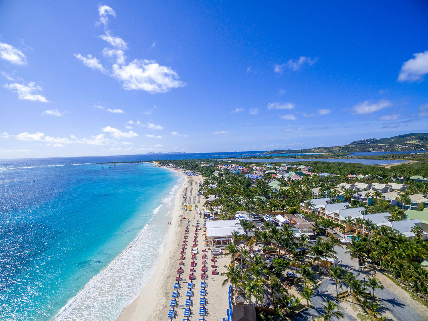 Aerial view of colourful sun loungers lined up on a white sand beach around a small tropical island 