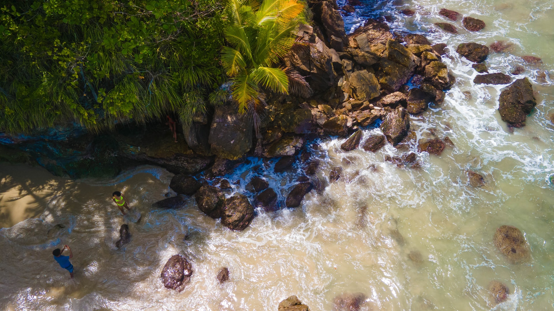 Aerial view of two people taking photos next to large rocks on a golden sand beach