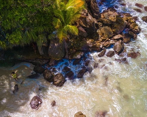 Aerial view of two people taking photos next to large rocks on a golden sand beach