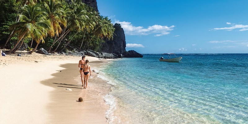 Two women walking along a white sand beach lined by tall palm trees and a mountain in the background