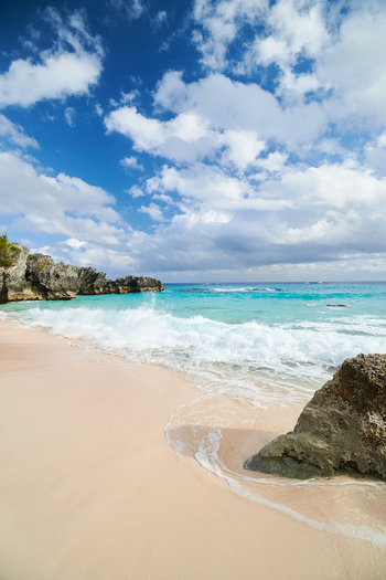 Beige sand beach with rocks in the background and on the right, fluffy waves and clouded sky