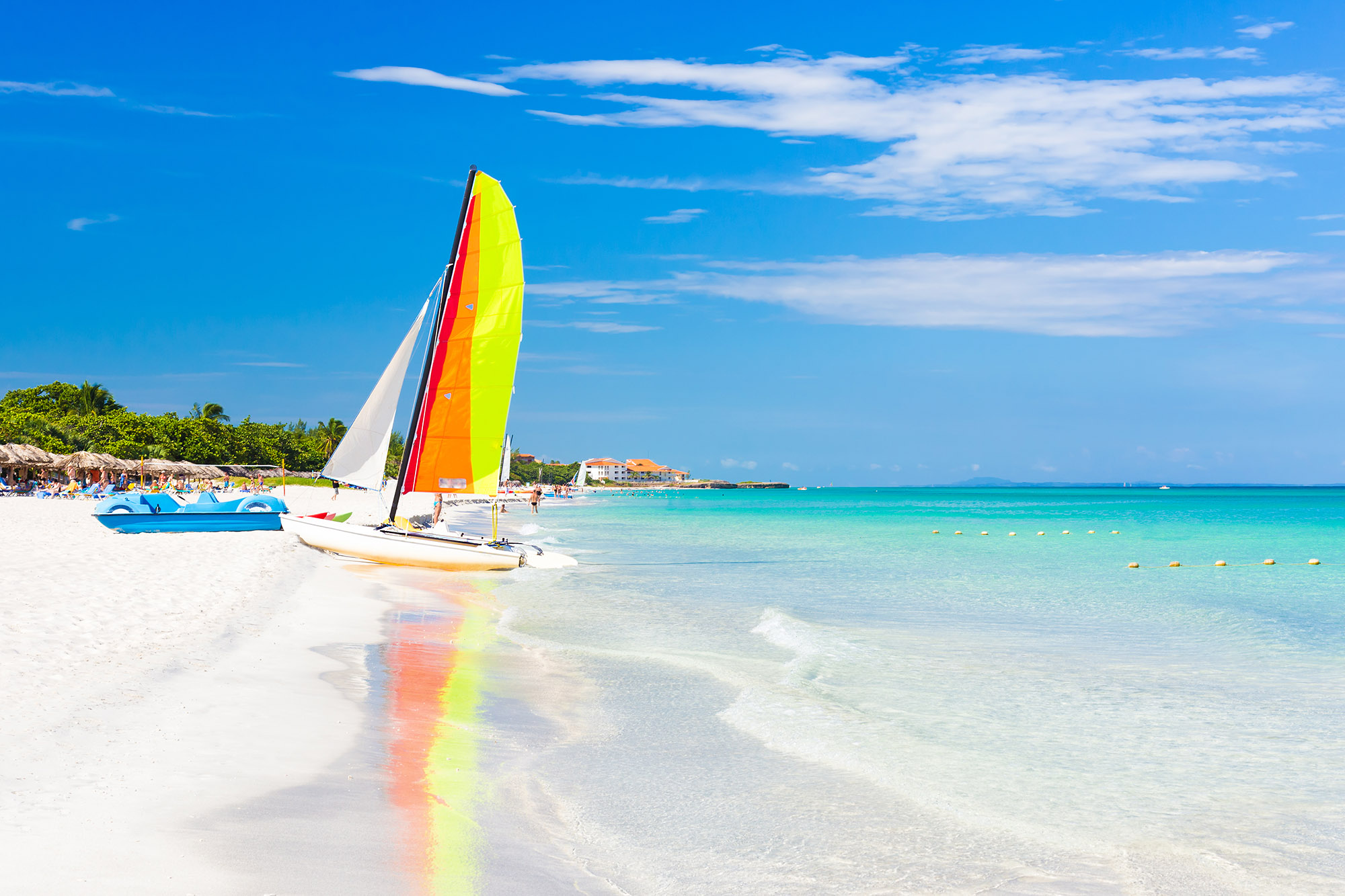 Sailing boat with colourful sail reflecting in the calm, clear blue sea next to a white sand beach 