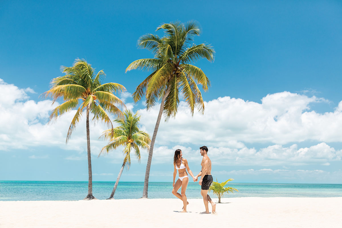 Young couple holding hands on white sand beach next to palm trees