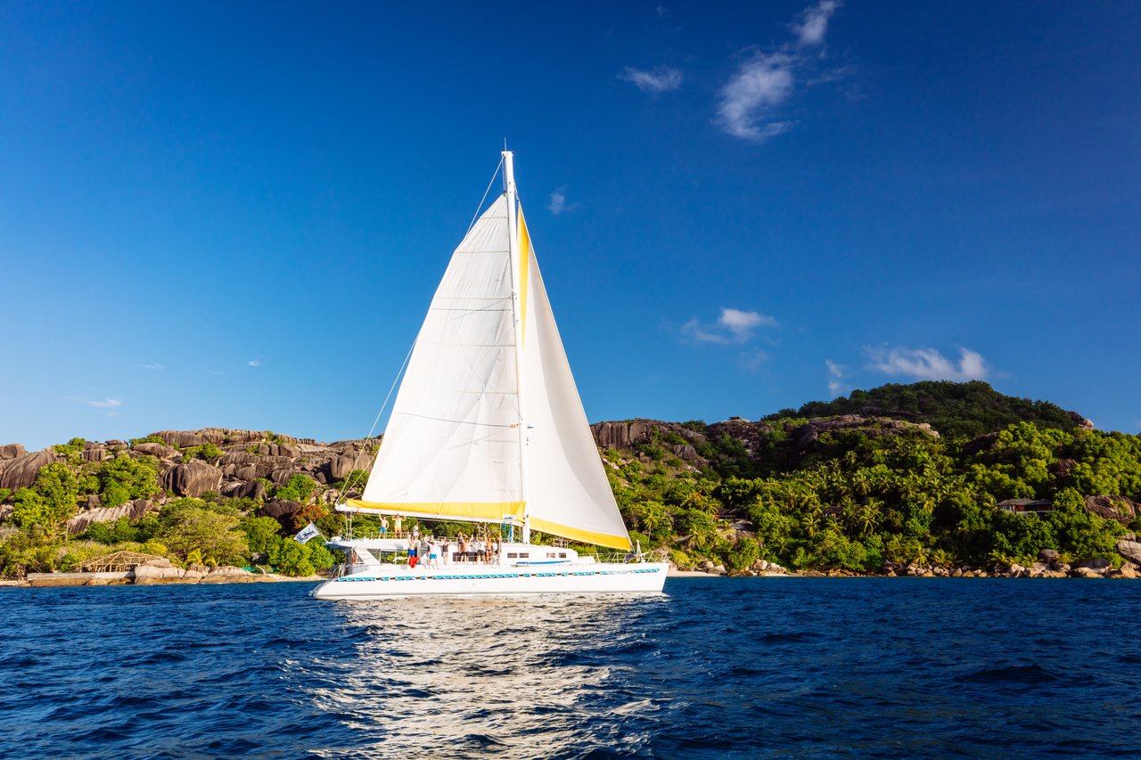 White sailing boat with large white sails on blue ocean with leafy green hills in background - Praslin 