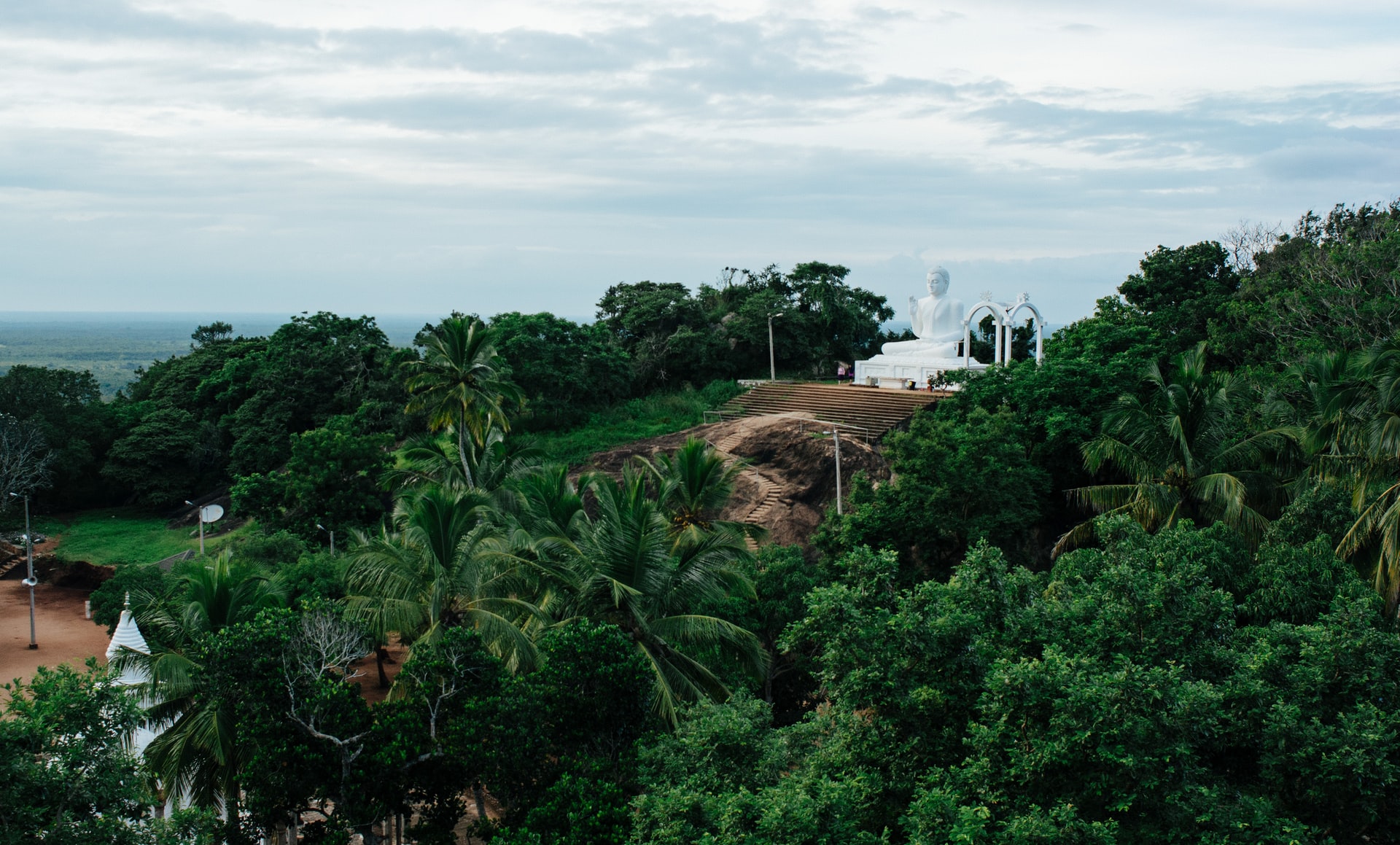 White Buddha statue on the top of a green hill covered in tropical trees 