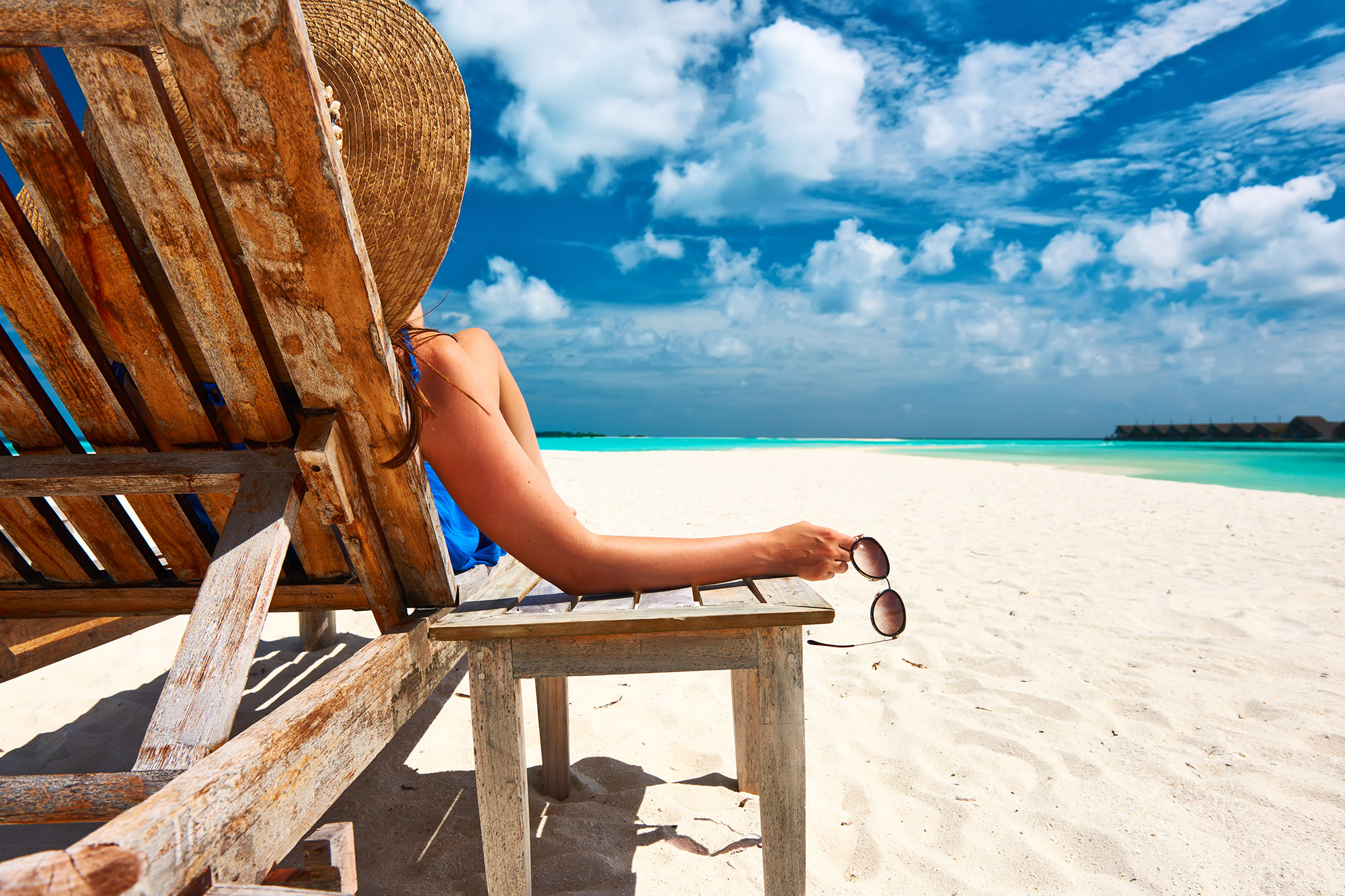 Woman holding a pair of sunglasses sitting on a wooden deck chair on a white sand beach
