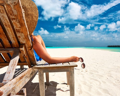 Woman holding a pair of sunglasses sitting on a wooden deck chair on a white sand beach