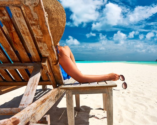 Woman holding a pair of sunglasses sitting on a wooden deck chair on a white sand beach