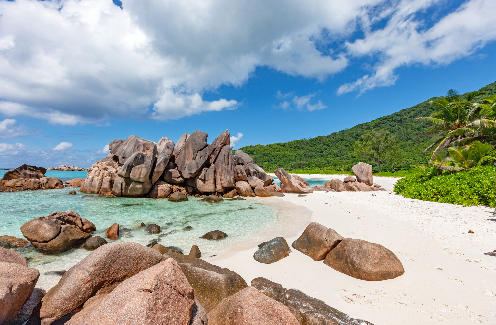 Granite boulders on white sand and crystal clear waters with hilly forest in background - Anse Cocos