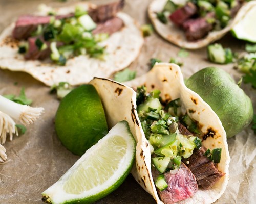 Meat tacos with a green relish placed next to sliced limes