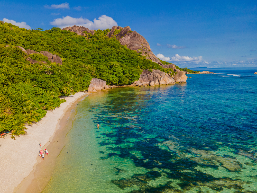 Turquoise ocean edging onto white sand with hilly green forest - Anse Source D'argent