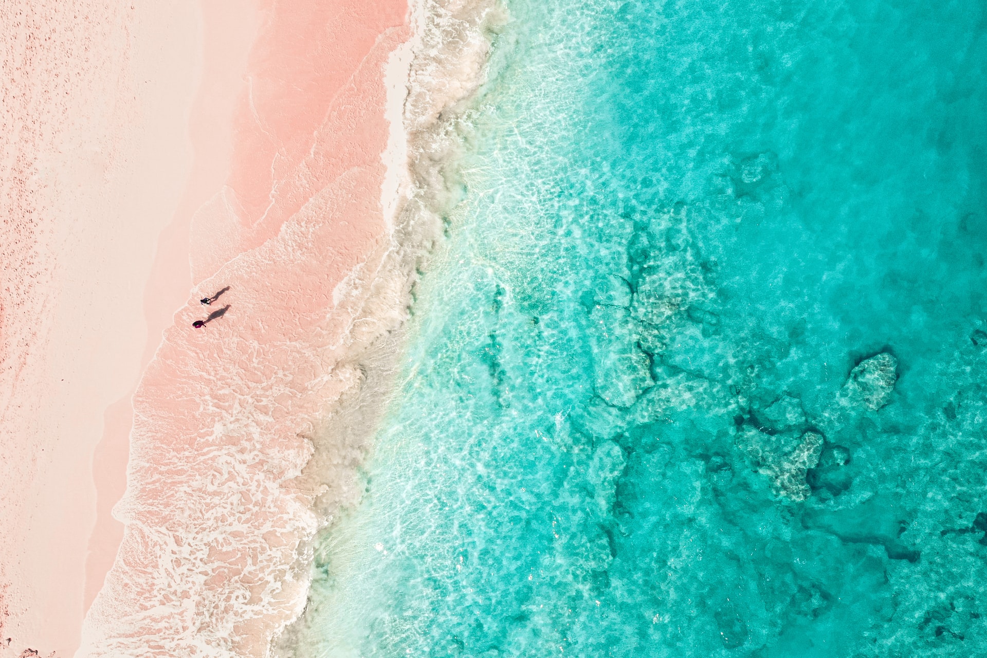 Aerial view of two people walking across a pink sand beach next to clear blue sea