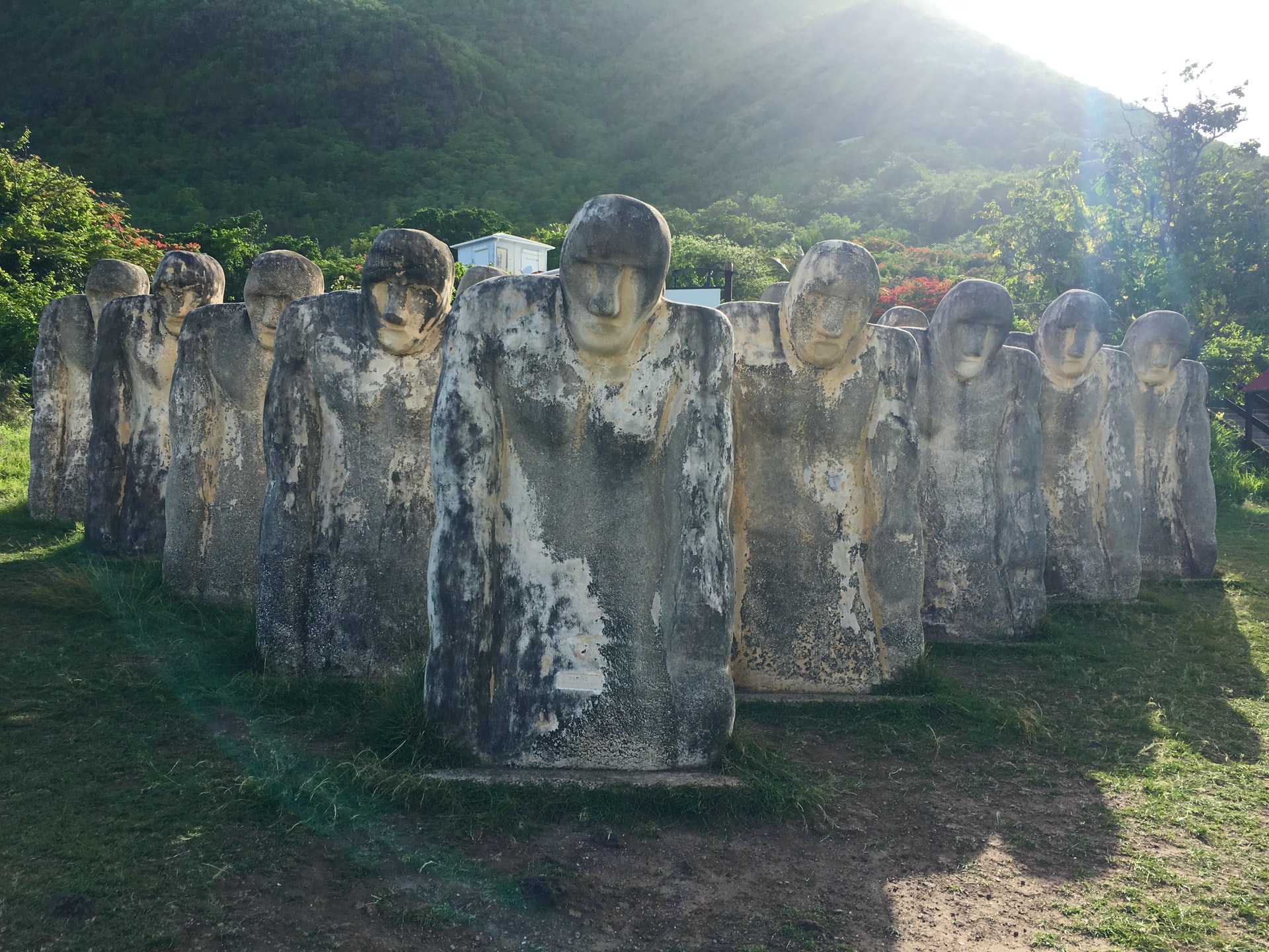 Sun shining on large stone statues of torsos at the bottom of a green mountai
