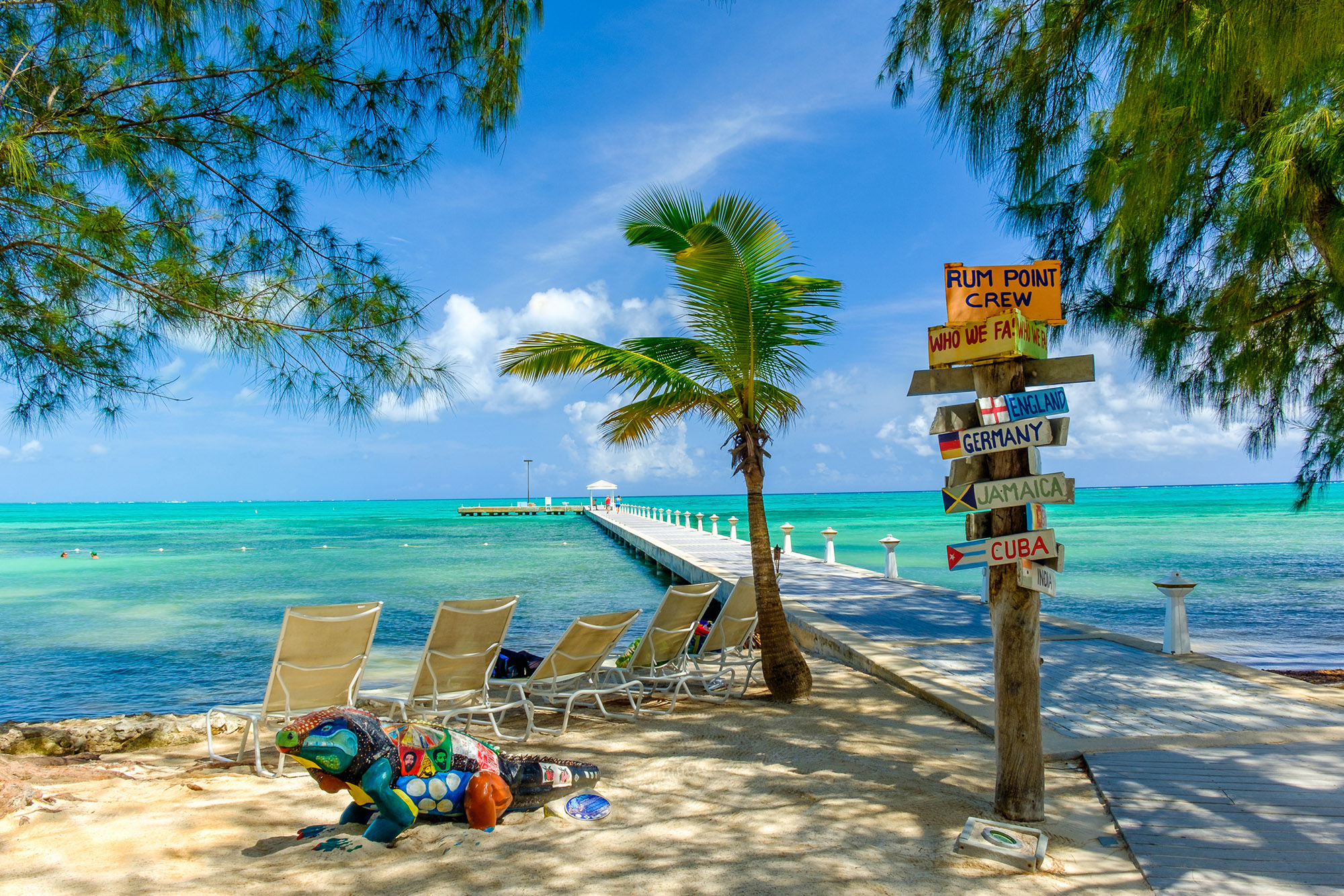 A colourful sign next to deckchairs laid out on a beach with a view of the bright blue sea - Rum Point Beach