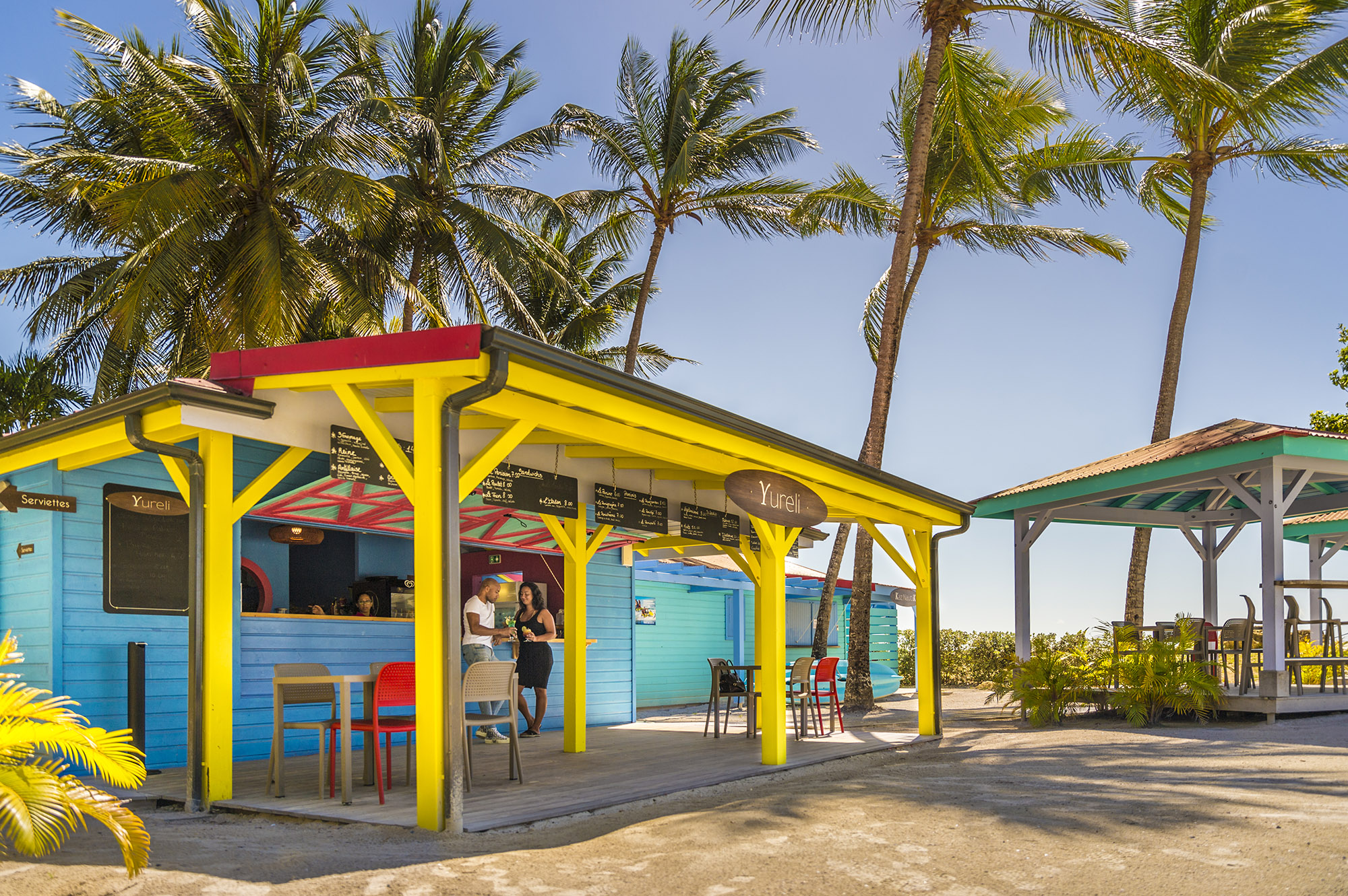 Man and woman holding drinks standing next to colourful beach shack with a sign that says 'Yureli'