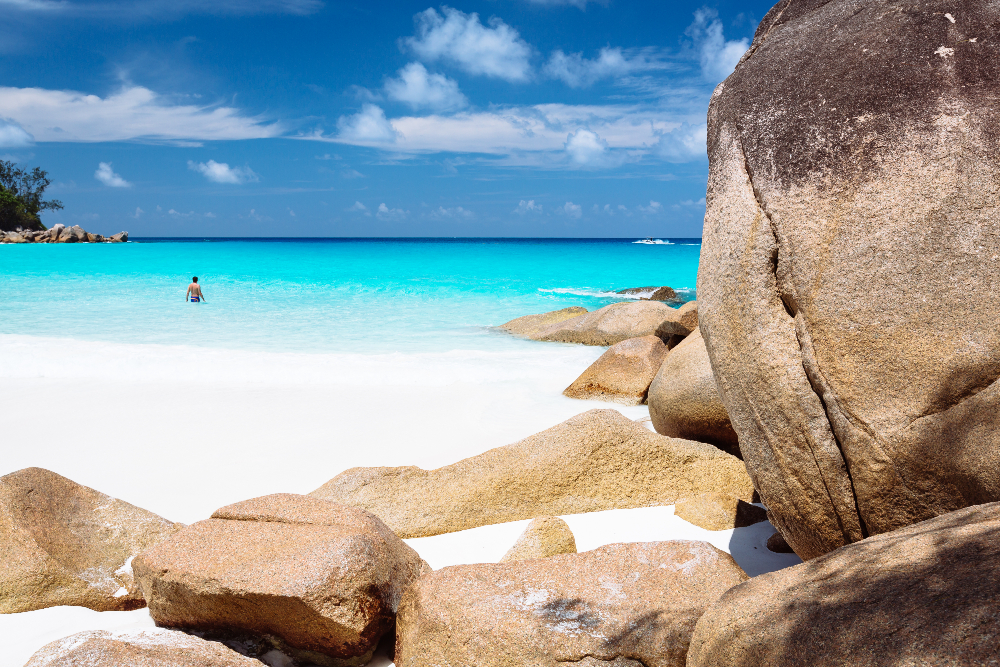 White sandy beach with lone man swimming in blue sea with rocks in foreground - Anse Georgette