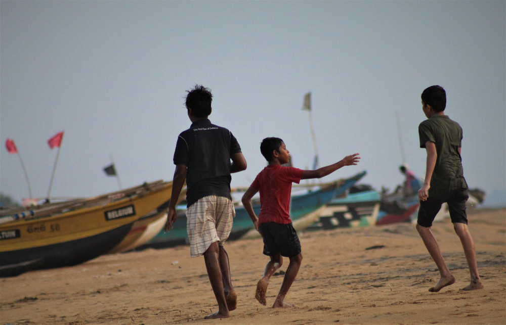 Three boys playing on beach with boats behind them at sunset - Wellawatte Beach 