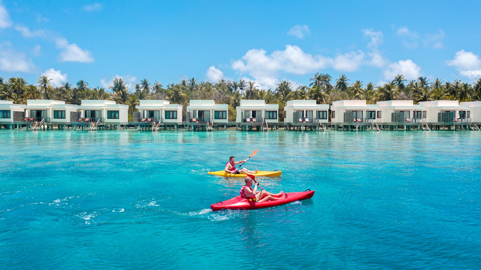 Two men in kayaks on bright blue sea going past overwater villas on a small island