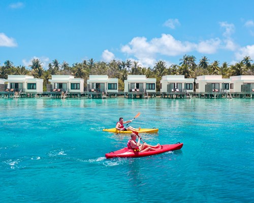 Two men in kayaks on bright blue sea going past overwater villas on a small island