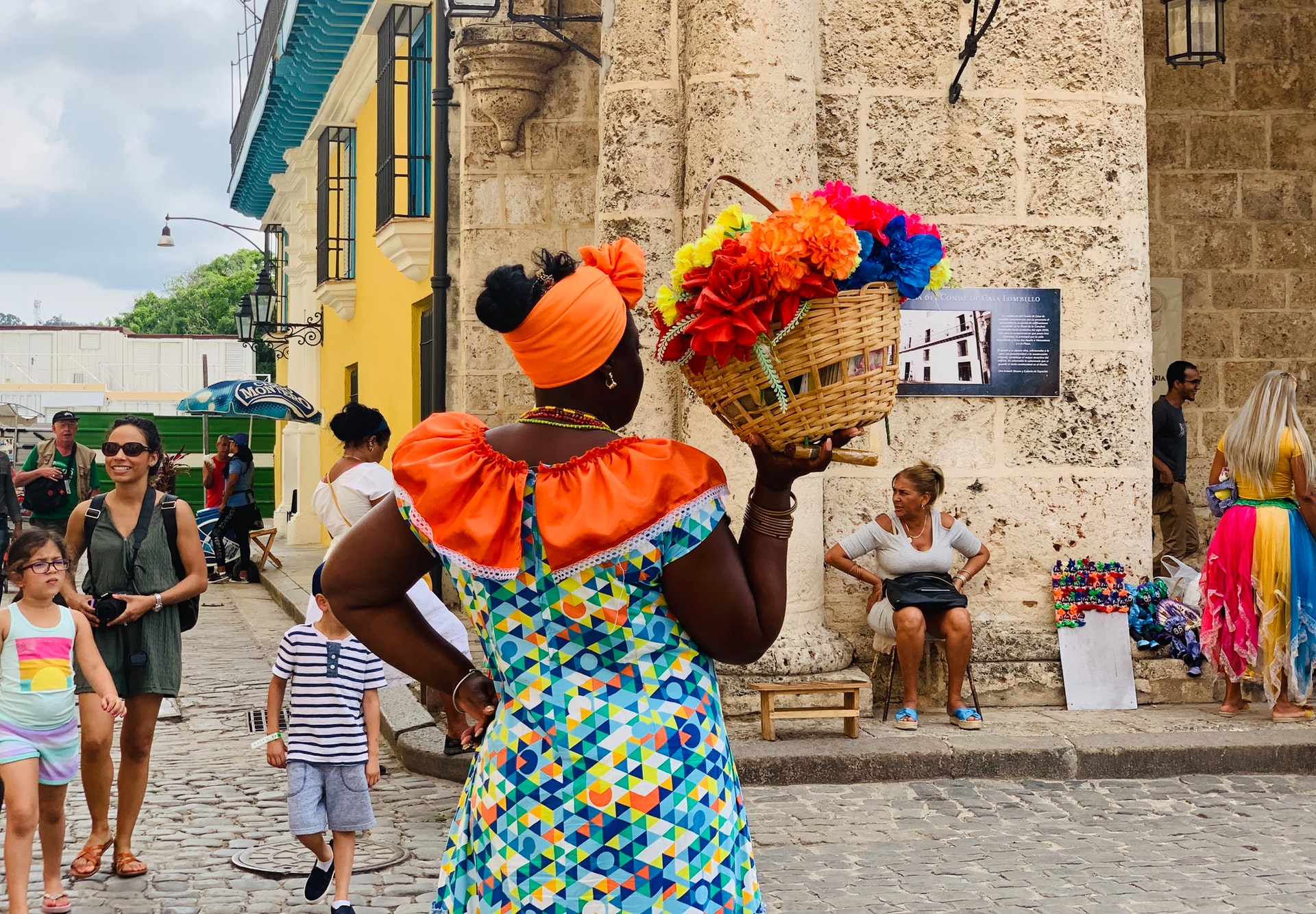 Standing woman in orange headdress carrying a straw basket of flowers through streets of Havana