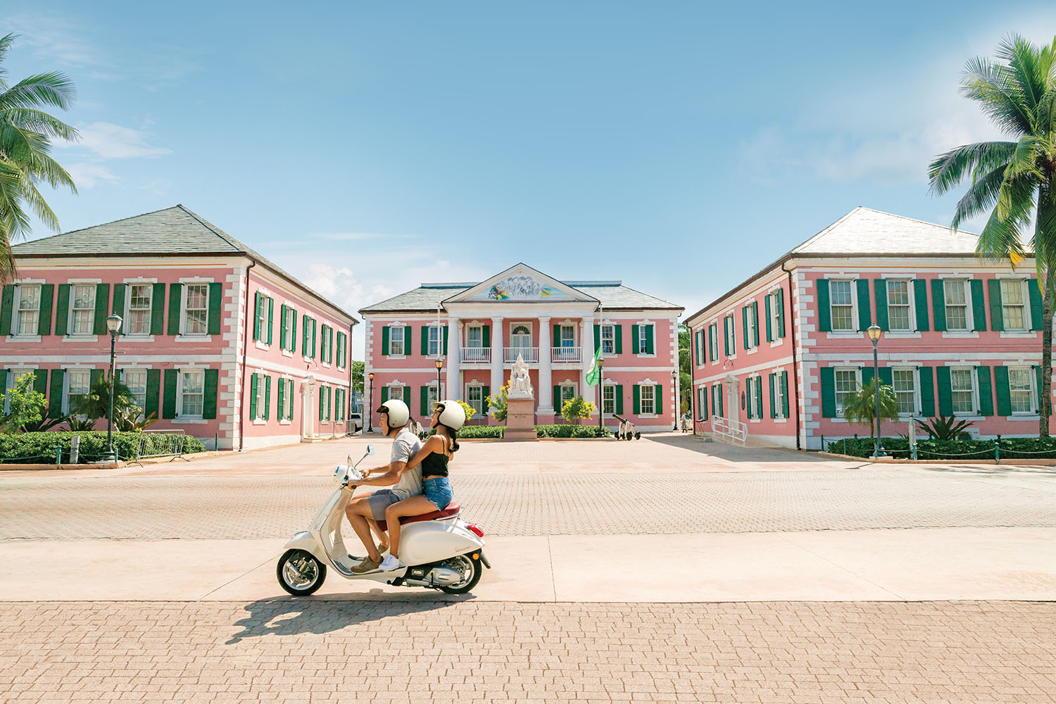 Couple on a scooter driving past several large pink buildings with green shutters