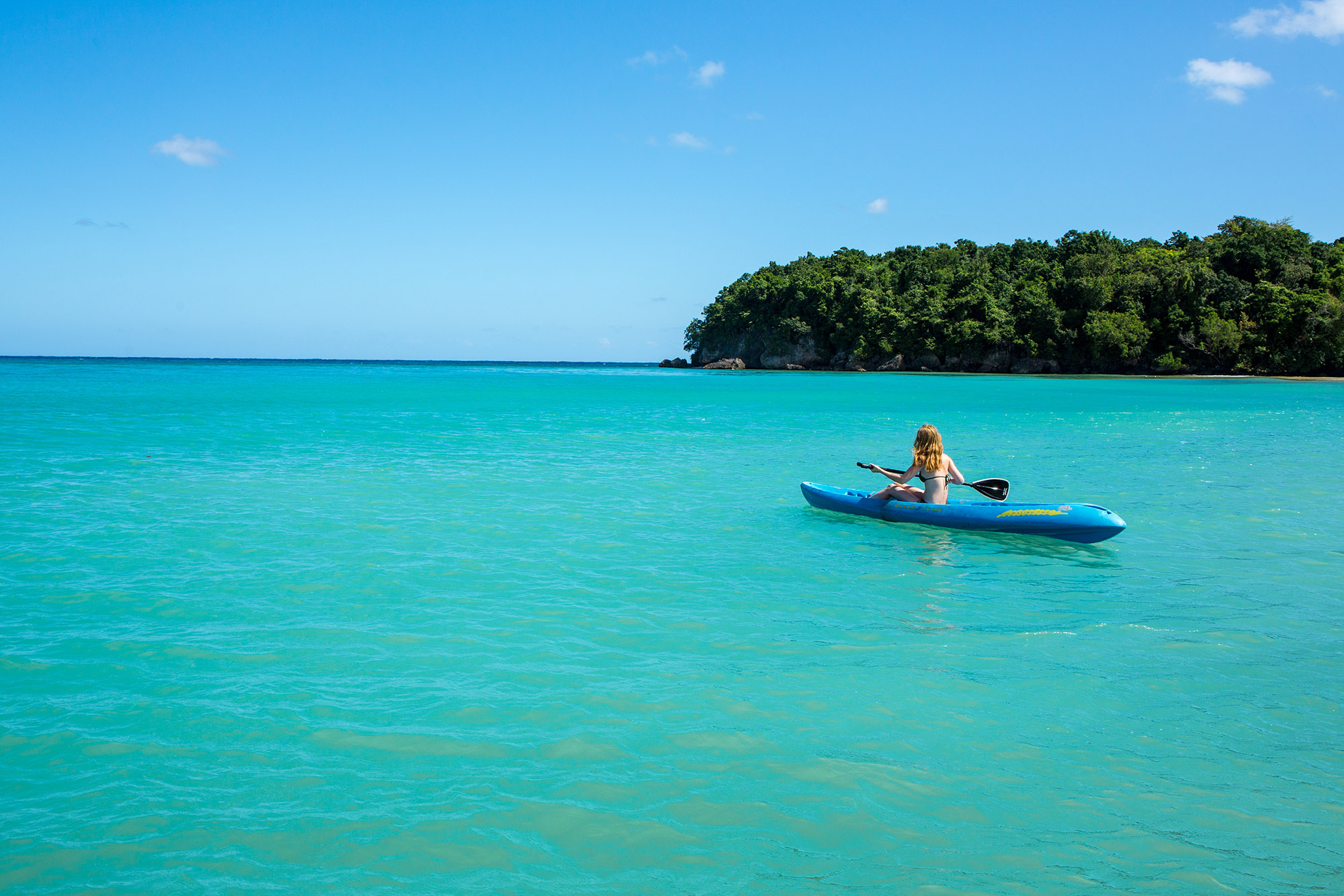Woman in blue kayak kayaking across a green lagoon with small forest-clad island in the background