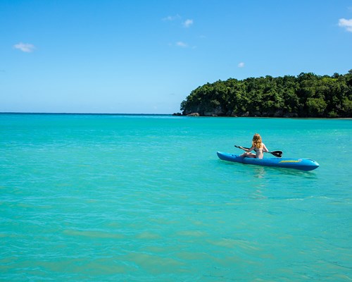 Woman in blue kayak kayaking across a green lagoon with small forest-clad island in the background