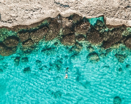 Aerial view of someone swimming in clear blue sea near land and rocks